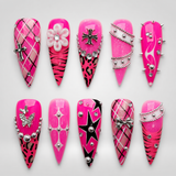 Ten coordinated hot-pink press-on nails displayed in two rows, featuring plaid, stripes, animal-inspired spots, stars, silver-toned charms, studs, and bead-chain accents.