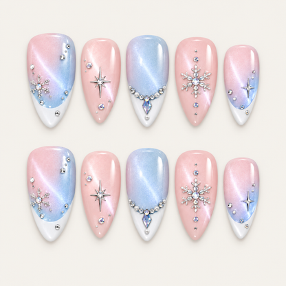 Ten blush pink and icy blue nails arranged in two rows on a greige background, featuring white tips, silver-toned snowflakes, starbursts, and jeweled accents.