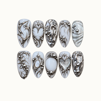 Ten milky white and silver nails arranged in two rows on a light background, featuring chrome swirls, starbursts, hearts, and bead-like accents.