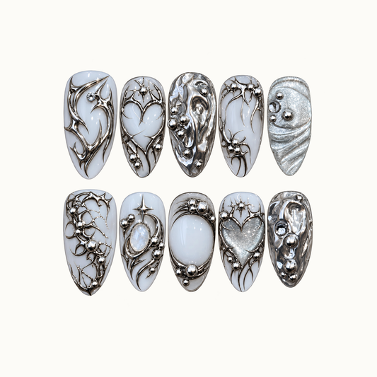 Ten milky white and silver nails arranged in two rows on a light background, featuring chrome swirls, starbursts, hearts, and bead-like accents.