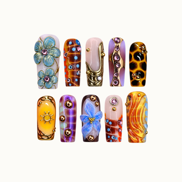 Ten decorated nails arranged in two rows on a pale background, featuring blue flowers, amber patterns, lavender accents, and gold-tone details.