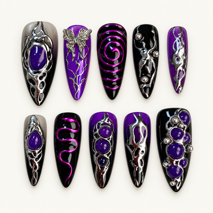 Ten glossy black and purple press-on nails with silver-tone ornate accents, butterfly art, violet rounded details, studs, and magenta swirl lines on a textured light background.