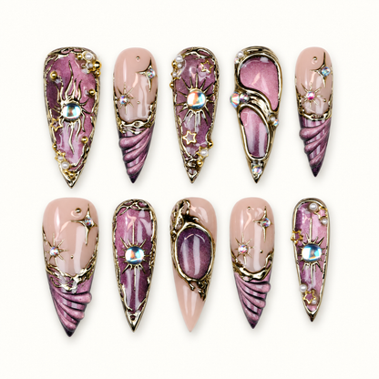 Ten coordinated mauve, plum, and nude-pink celestial press-on nails with gold-tone orbit frames, sunbursts, stars, pearls, and crystal accents.