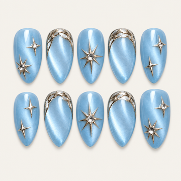 Ten pearly blue press-on nails arranged in two rows on a warm neutral background, with metallic-looking stars and looping accents.