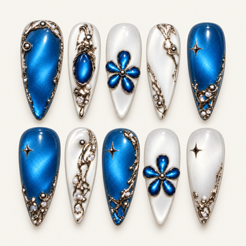 Catalog view of ten blue and pearly white press-on nails with raised blue flowers, star accents, and metallic-toned scrollwork on a warm light background.