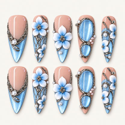 Ten blush pink and shimmering blue press-on nails arranged in two rows, with blue-and-white flowers, oval blue accents, silver-toned flourishes, and pearl-like beads.