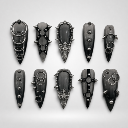 Ten glossy charcoal-black gothic press-on nails arranged in two rows, featuring silver-toned rings, studs, spikes, and ornate raised details.