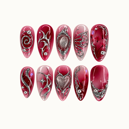 Ten crimson and blush nails arranged in two rows on a light neutral background, with silver-tone swirls, butterfly-wing outlines, and shimmering heart and oval accents.