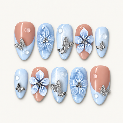 Ten blue and peach press-on nails arranged in two rows, with raised blue flowers, silver butterflies, white dots, and a glossy finish.
