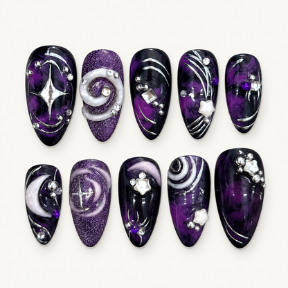 Ten coordinated glossy press-on nails in black and deep purple with silver orbit lines, crystals, glitter, and raised spiral accents displayed in a clear case.