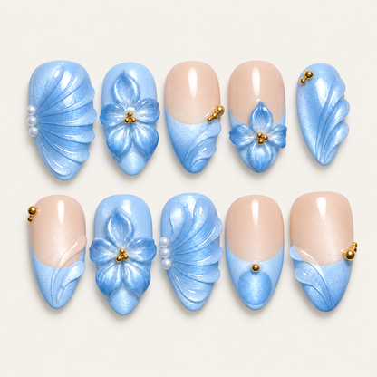 Ten pale blue and nude press-on nails arranged in two rows, with blue flowers, fan-like curves, pearl-like accents, and gold-tone beads on a light background.