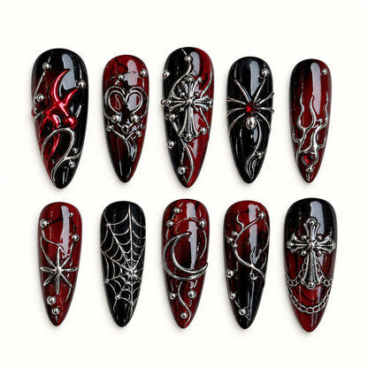 Ten coordinated glossy black and deep red press-on nails with raised silver-toned spiderweb, spider, cross, crescent, and starburst details on a light neutral background.