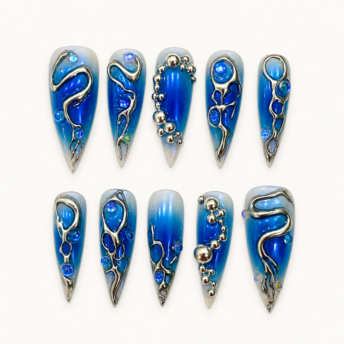 Ten blue chrome press-on nails arranged on a light textured background, detailed with silver flowing lines, oval accents, and metallic bead clusters.
