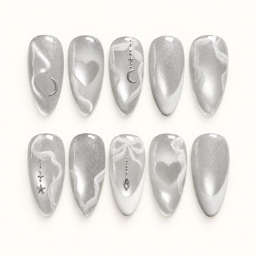Ten silver shimmer nails in two rows on a pale background, with white swirls, hearts, crescent moons, stars, and a bow accent.