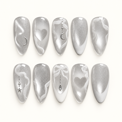 Ten silver shimmer nails in two rows on a pale background, with white swirls, hearts, crescent moons, stars, and a bow accent.