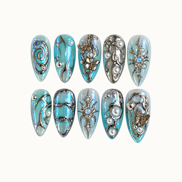 Ten icy blue and aqua nails in two rows on a light neutral background, decorated with silver-tone swirls, butterflies, starbursts, and pearl-like accents.