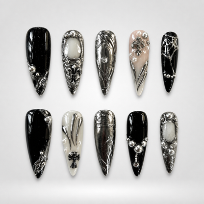 Ten coordinated gothic press-on nails in glossy black, ivory, blush nude, and reflective silver with ornate crosses, beads, and metallic details.
