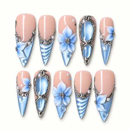 Ten coordinated press-on nails arranged in two rows on a light background, showing pink bases, blue pointed tips, blue flowers, silver-tone scrollwork, and pearl-like accents.