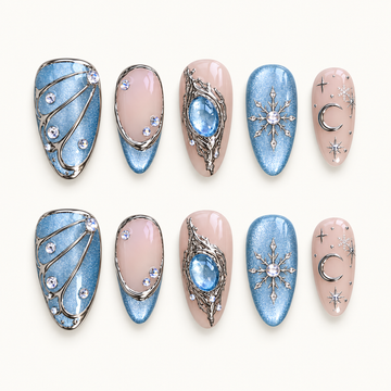 Ten Moonlit Frost press-on nails arranged in two rows, showing icy blue shimmer, blush pink, silver-tone accents, snowflakes, a crescent moon, and a blue centerpiece.