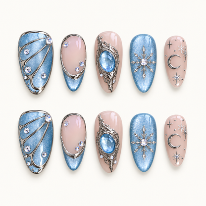 Ten Moonlit Frost press-on nails arranged in two rows, showing icy blue shimmer, blush pink, silver-tone accents, snowflakes, a crescent moon, and a blue centerpiece.