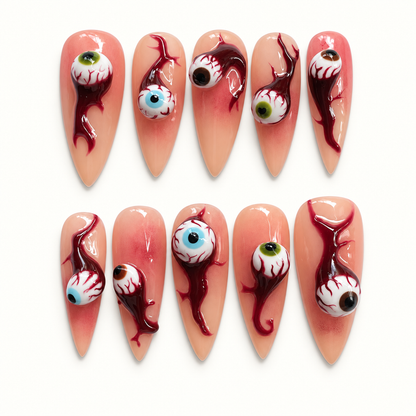 Ten glossy nude-peach press-on nails arranged on a pale background, featuring raised bloodshot eyeballs and deep burgundy drip artwork.