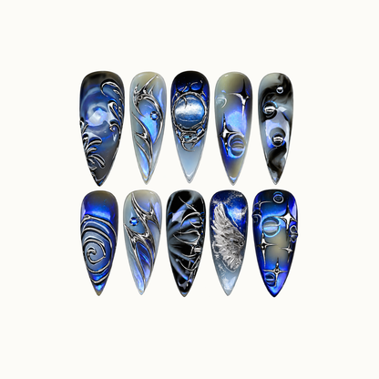 Ten pointed nails arranged in two rows on a light neutral background, featuring cobalt blue, smoky gray, black, and silver celestial artwork.