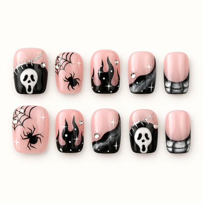 Ten coordinated blush pink and black press-on nails with spiderwebs, spiders, smoky gray details, abstract face motifs, and rhinestone-style accents on a pale neutral background.