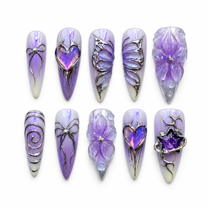Ten Lavender Orbit press-on nails arranged on a pale background, showing violet marbling, raised petals, silver-toned linework, hearts, and celestial-inspired accents.
