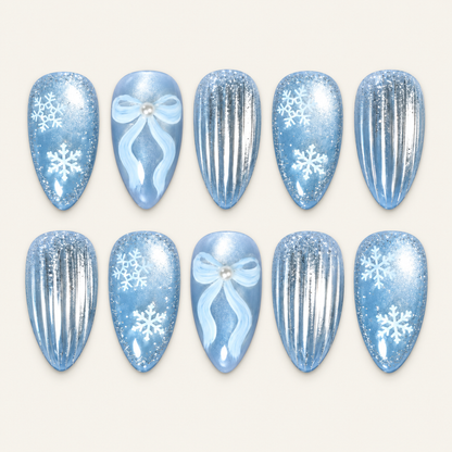 Ten icy blue press-on nails arranged in two rows on a light greige background, featuring white snowflakes, pale blue bows, and silver vertical stripes.