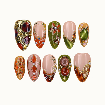 Ten ornate nails arranged in two rows on a pale background, featuring olive, blush, and amber colors with gold-tone botanical and jewel-like details.