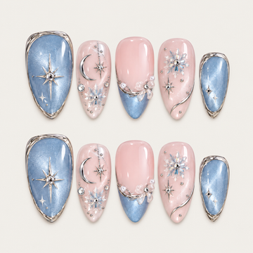 Ten blush pink and shimmering blue nails arranged in two rows on a greige background, with silver-tone moons, stars, snowflakes, and bow accents.