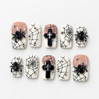 Ten coordinated Eternal Web press-on nails arranged in a clear case, with black spiderwebs, raised spiders, black crosses, ivory and blush accents, and metallic studs.