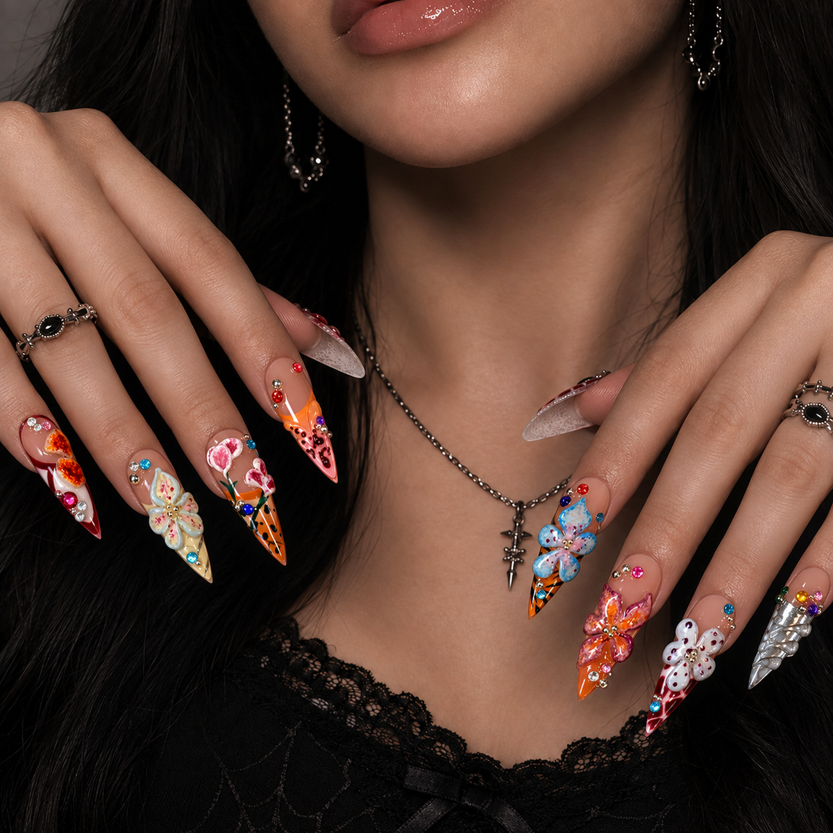 Hand model mockup showing pointed floral nails with raised blooms, colorful gems, and contrasting orange, burgundy, pink, and silver accents.