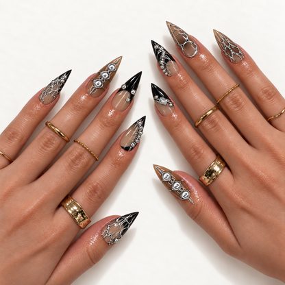 Two hands model pointed taupe and black nails with silver-tone looping lines, thorn-like flourishes, rounded accents, and a rectangular detail.