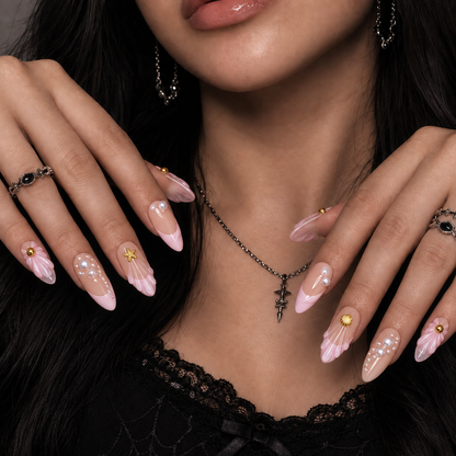 Model wearing blush and pink shell-inspired nails with pearl-like accents and gold-tone charms, with both hands posed near the neckline.