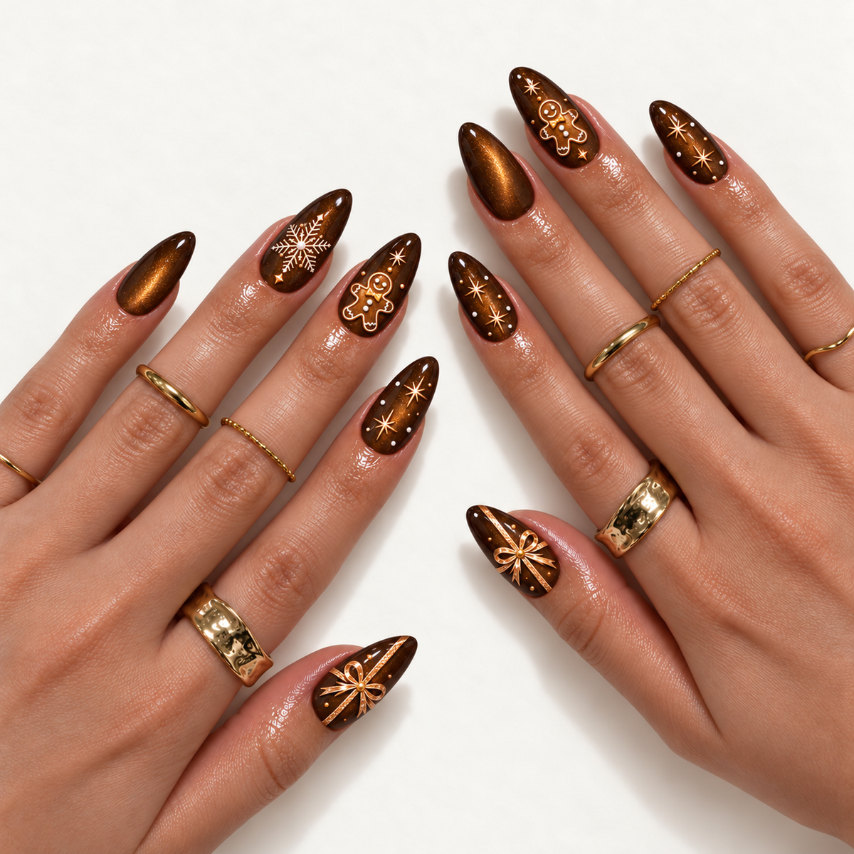 Two hands modeling pointed brown and amber shimmer nails with gingerbread figures, white snowflakes, gold-toned bows, and star accents, alongside gold-toned rings.