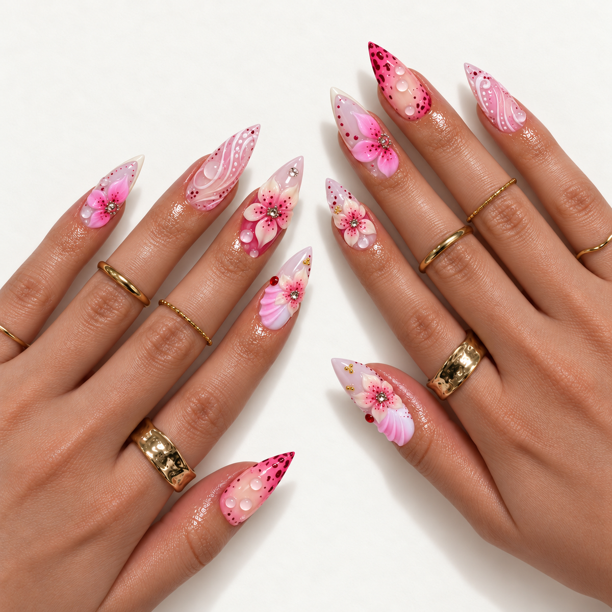 Two hands wearing pointed pink floral nails with raised petals, sparkling centers, clear droplets, and white swirls, styled with gold-toned rings.