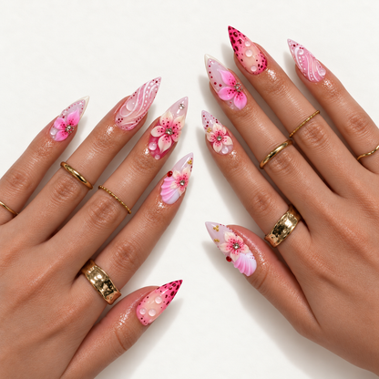 Two hands wearing pointed pink floral nails with raised petals, sparkling centers, clear droplets, and white swirls, styled with gold-toned rings.