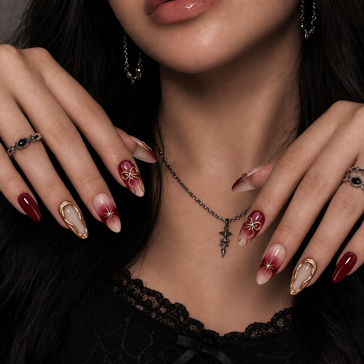 Hands modeling burgundy and blush nude nails with gold bows, starbursts, and gold border accents against black lace clothing.