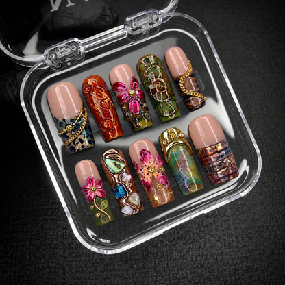 Open clear case holding ten decorated nails on dark textured fabric, showing floral motifs, olive mosaics, amber swirls, and gold-tone details.