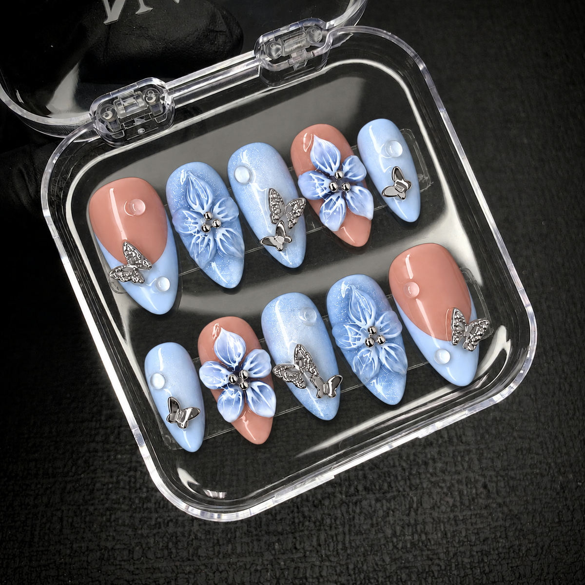Open clear case displaying ten blue and peach nails with raised flowers, silver butterflies, and white dot accents.