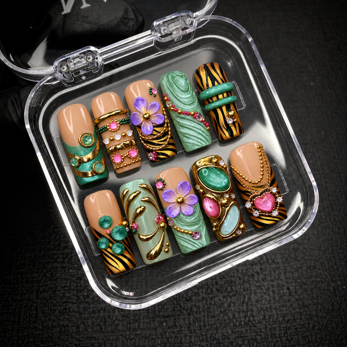 Open clear nail case on a dark textured background holding ten coordinated nails with lavender flowers, green accents, gold-tone chains, and a pink heart.