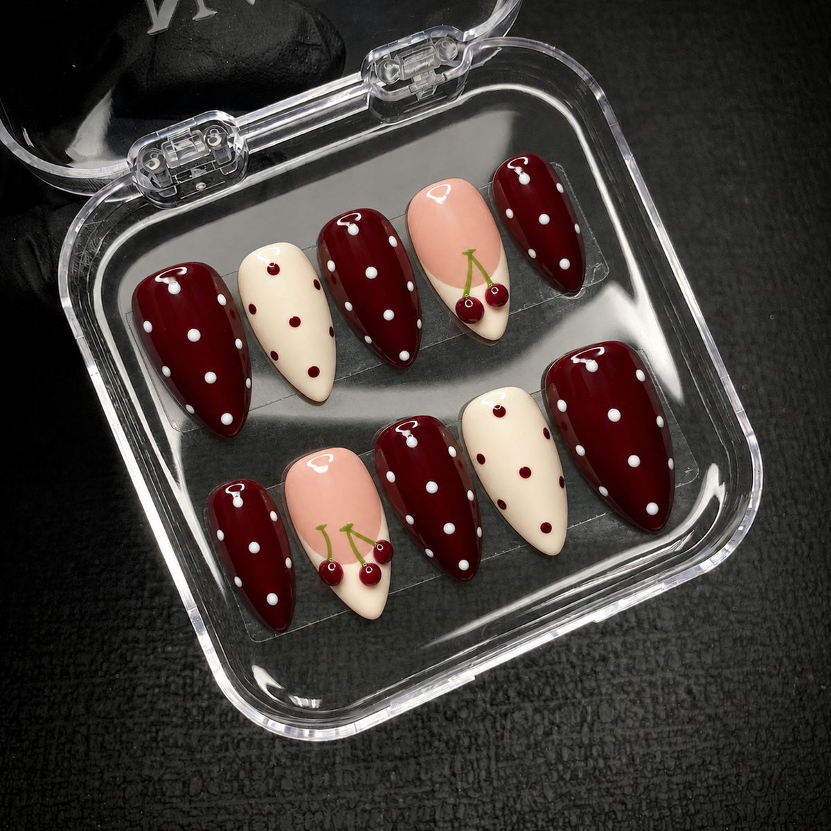 Open clear case on a dark textured surface holding ten coordinated burgundy, ivory, and blush pink nails with polka dots and cherries.