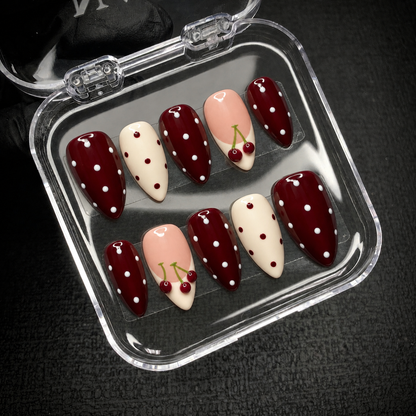 Open clear case on a dark textured surface holding ten coordinated burgundy, ivory, and blush pink nails with polka dots and cherries.
