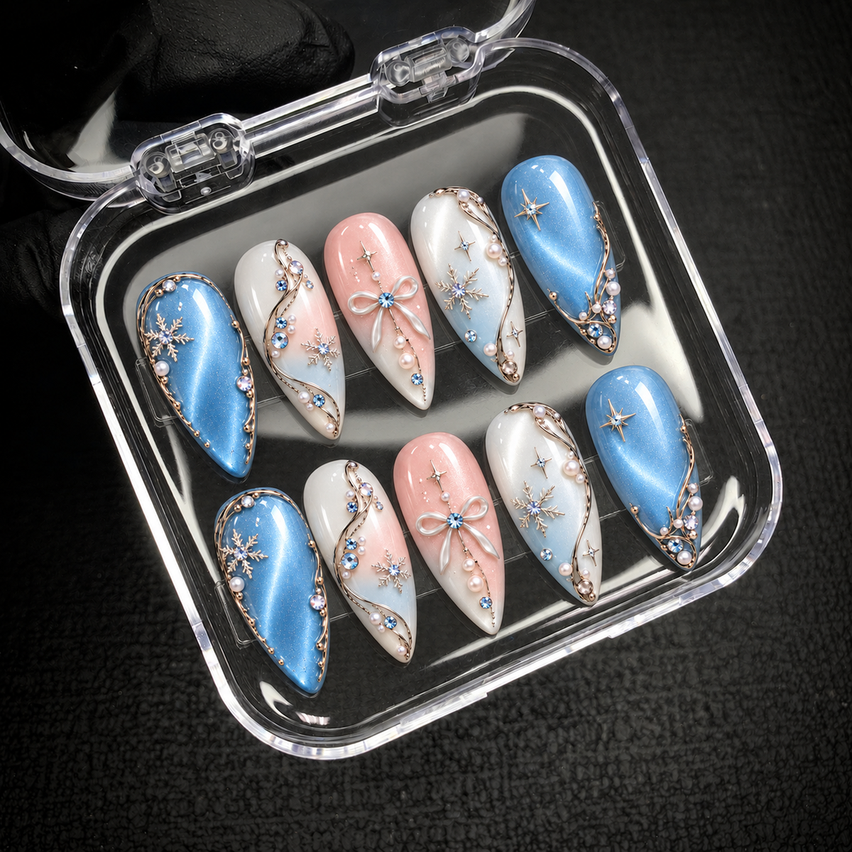 Open clear nail case on a dark textured surface holding ten blue, pink, and white nails with winter motifs and dimensional accents.