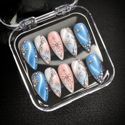 Open clear nail case on a dark textured surface holding ten blue, pink, and white nails with winter motifs and dimensional accents.
