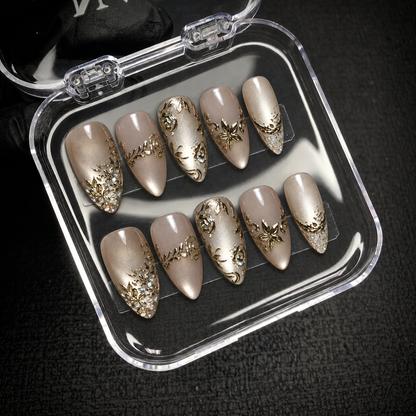 Open clear nail case on a dark textured surface holding ten champagne and greige nails with gold-toned floral details.