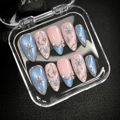 Open clear nail case on dark textured fabric holding ten coordinated pink and blue nails with celestial motifs and silver-tone accents.