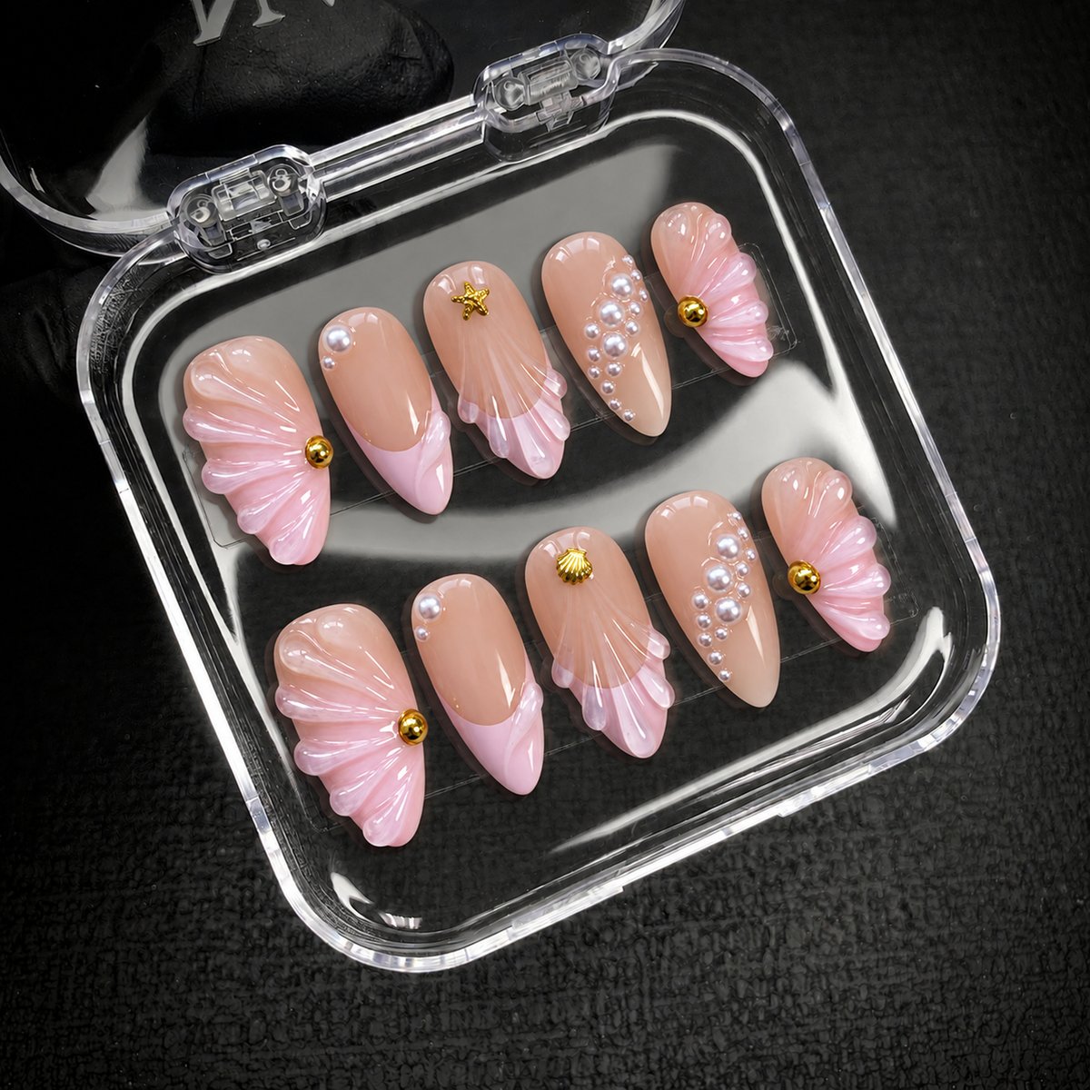 Open clear display box on a dark textured surface holding ten blush nails with sculpted pink shell details, pearl-like accents, and gold-tone motifs.