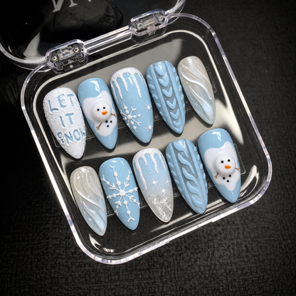Open clear case holding ten coordinated icy blue and white winter nails with snowmen, snowflakes, textured lettering, and sweater-inspired accents.